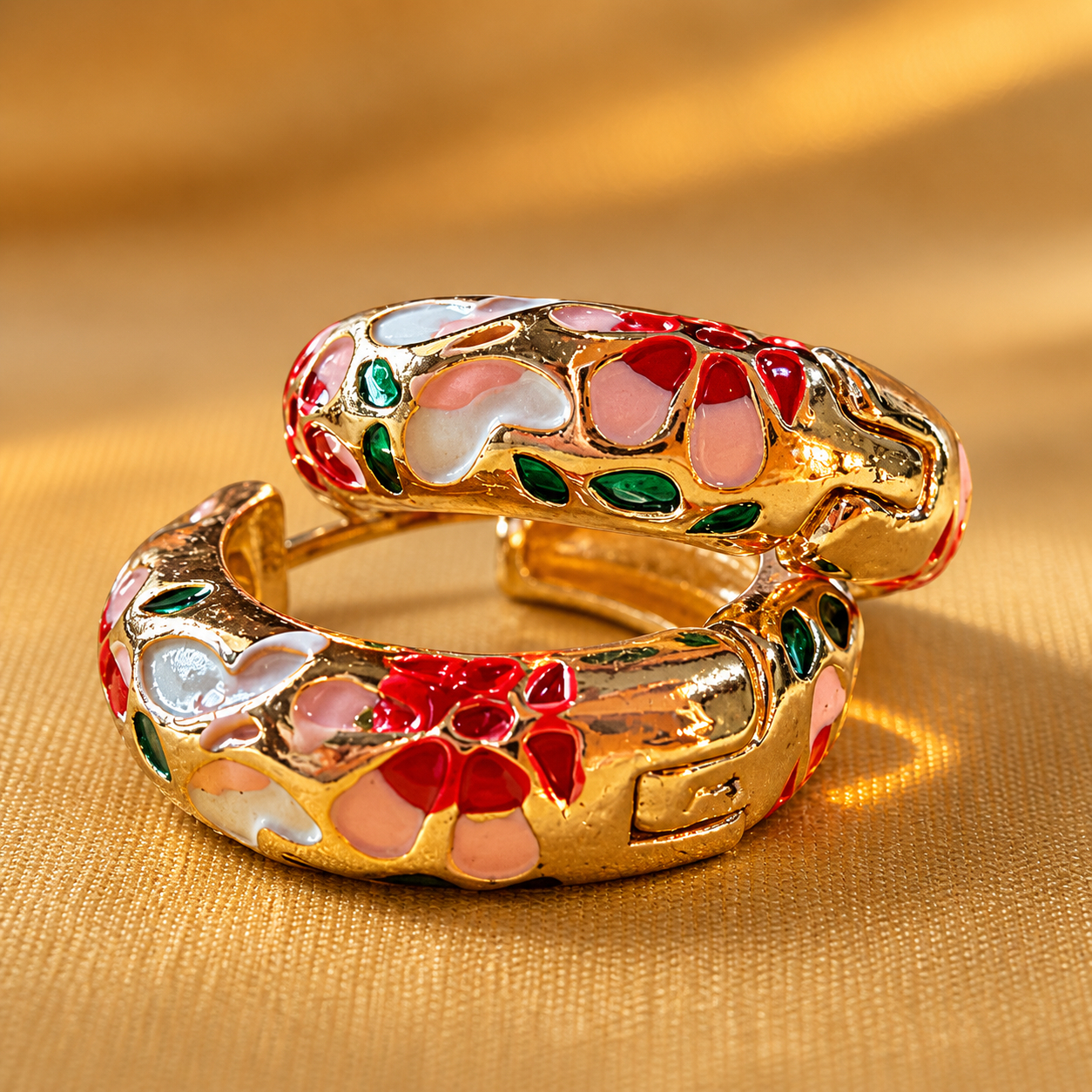 Pair of gold-plated enamel hoop earrings with red, pink, white, and green floral design on a warm beige linen background — House of Ruhira