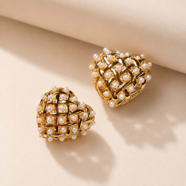 Gold-toned heart-shaped stud earrings with woven lattice and faux pearls on a warm beige linen background — House of Ruhira