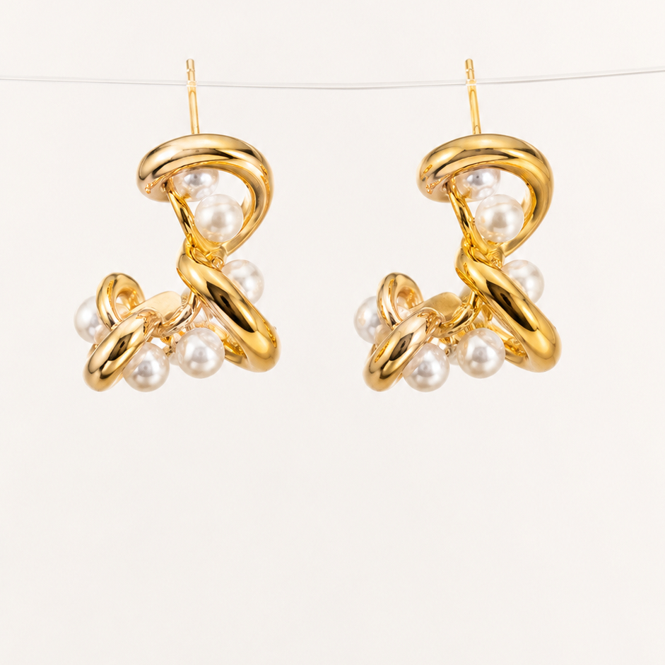 A pair of elegant gold pearl drop earrings featuring intertwined metal and white pearls, resting on a warm beige linen fabric — House of Ruhira