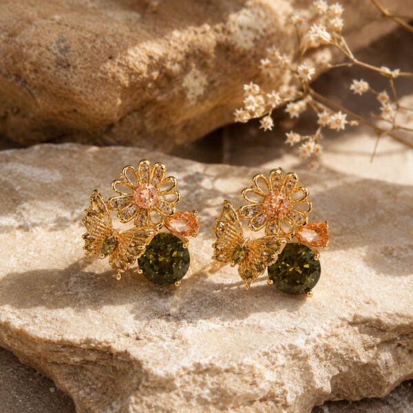 Gold-plated floral and butterfly stud earrings with olive green, peach, and clear cubic zirconia on a warm beige linen background — House of Ruhira
